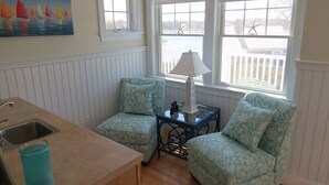 Una habitación acogedora con kitchenette, dos sillones estampados, una mesita con lámpara y un cuadro con barcos a vela en la pared.