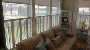 Un salón acogedor con un sofá a cuadros, una mesa de centro de vidrio y vistas a un lago a través de grandes ventanas con persianas.