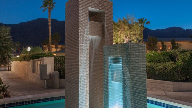 A pool with a waterfall feature, surrounded by a brick wall and palm trees.