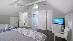 A bedroom with two beds, a ceiling fan, a television, and a bathroom visible through an open door. - Ride the Tide – Heated Pool Home | Steps to  Holmes Beach | Heated Pool | Free WIFI & Trolley , NO Roads to Cross, Dog*(See Note)Allow (Holmes Beach)