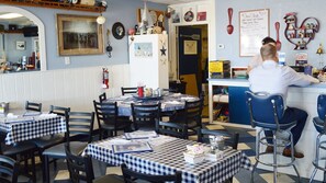 A restaurant interior with checkered tablecloths, black chairs, a counter with a person serving, and nautical-themed decorations.