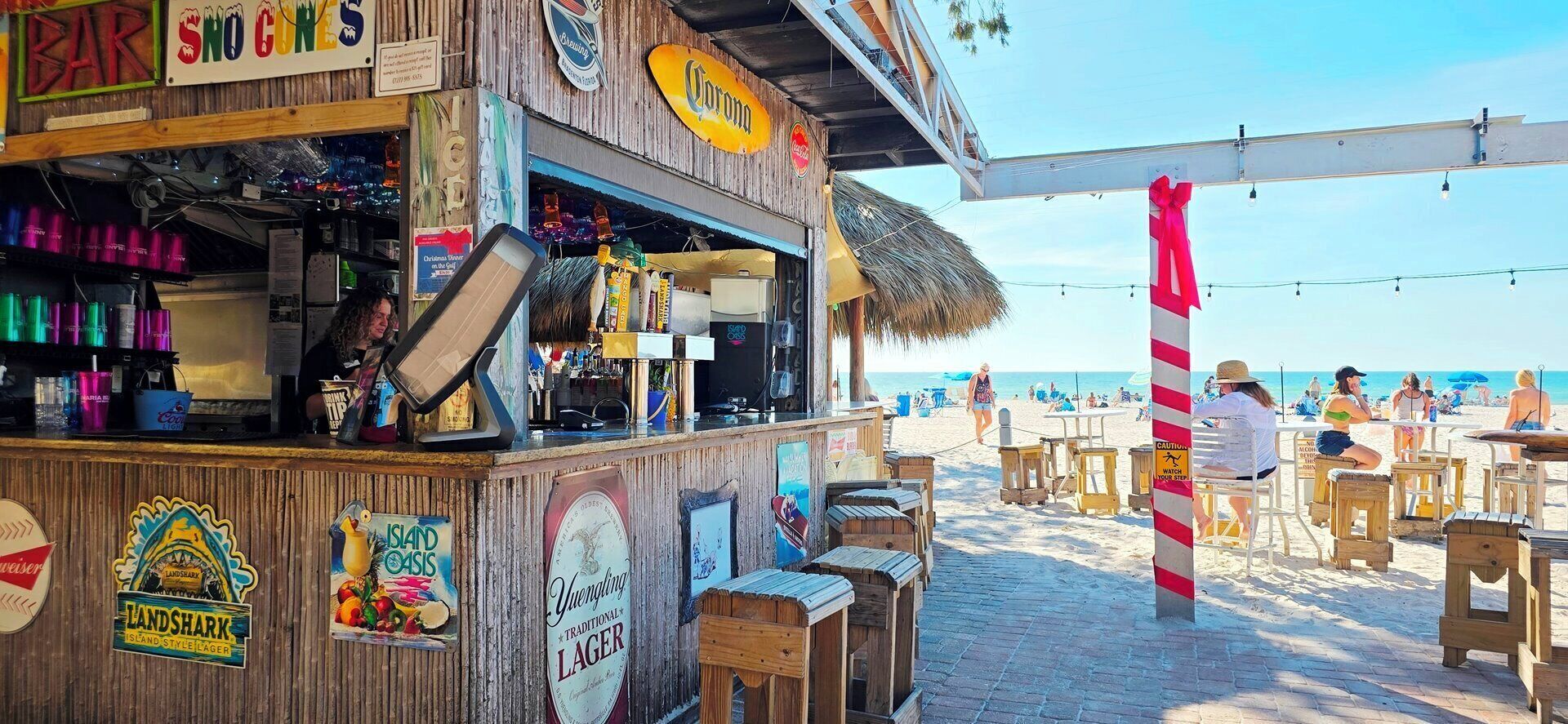 Eine Bar am Strand mit Holzbauten, einem Schild für 'Landshark Island Style Lager' und Blick auf den Strand mit Menschen im Hintergrund.
