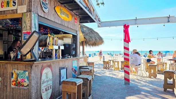 Un bar situé en bord de mer, avec des structures en bois, une enseigne indiquant « Landshark Island Style Lager », et une vue sur la plage avec des personnes en arrière-plan.