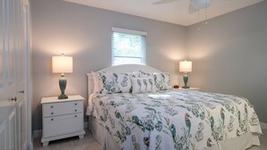 Une chambre à coucher comprenant un lit, deux tables de chevet, une commode, une fenêtre avec stores et un ventilateur de plafond.