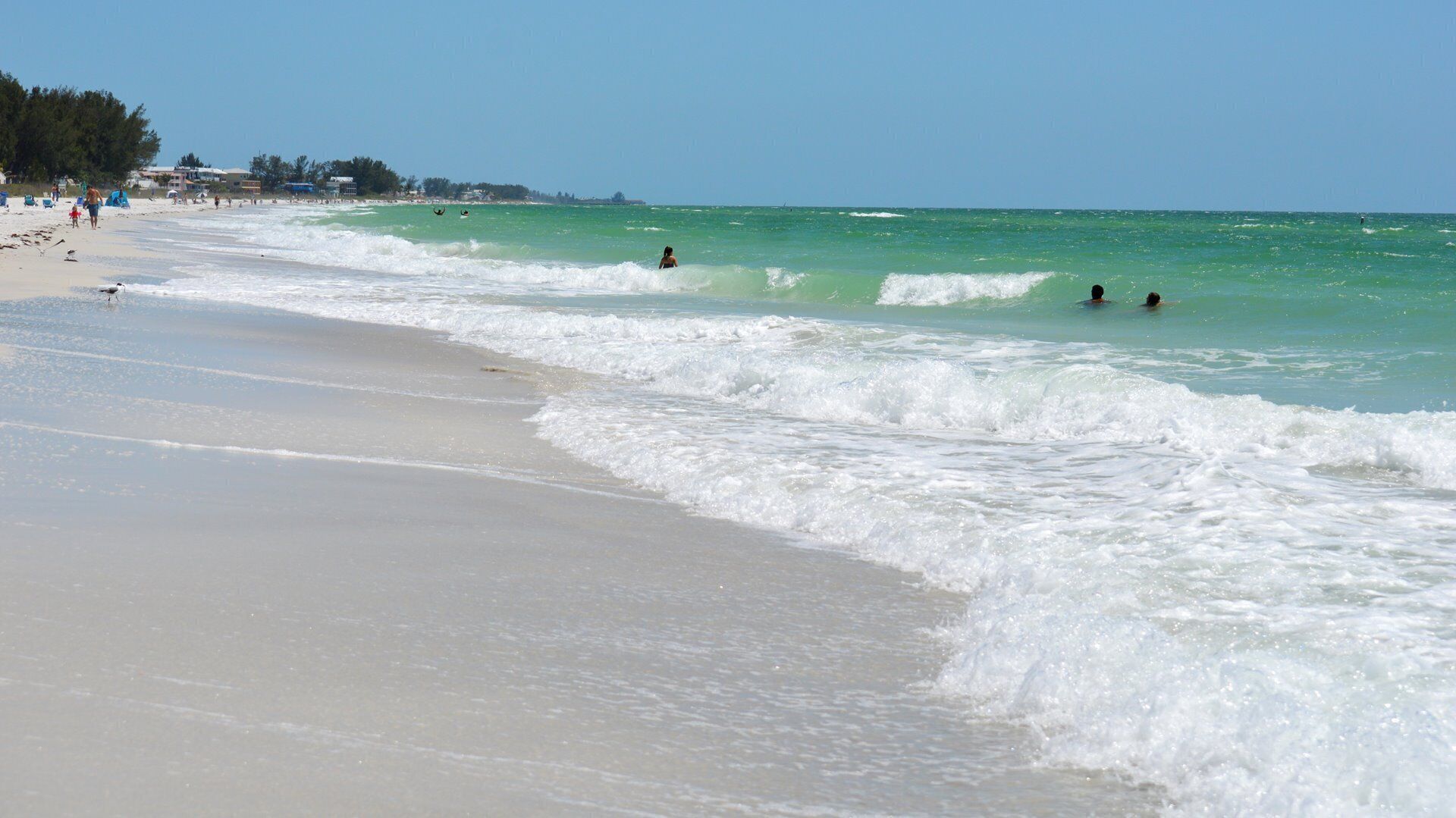 A beach with waves, people, and buildings in the distance.