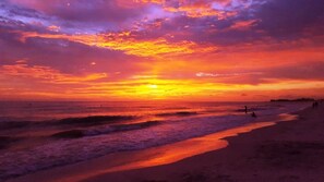 A beach scene with waves, a sunset, and a few people on the shore. - Ride the Tide – Heated Pool Home | Steps to  Holmes Beach | Heated Pool | Free WIFI & Trolley , NO Roads to Cross, Dog*(See Note)Allow (Holmes Beach)