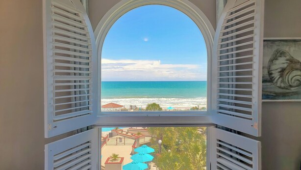 Blick durch ein Fenster mit Rollläden auf einen Strand und das Meer.