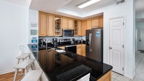 A modern kitchen with wooden cabinets, a black countertop, stainless steel appliances, and a white door. - GD 504 Emerald Sea Oasis: Wake Up to Breathtaking Ocean Views (Fort Walton Beach)