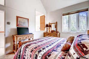 Un dormitorio con una cama grande, un tocador, un televisor, un espejo y una ventana con vistas a un bosque.
