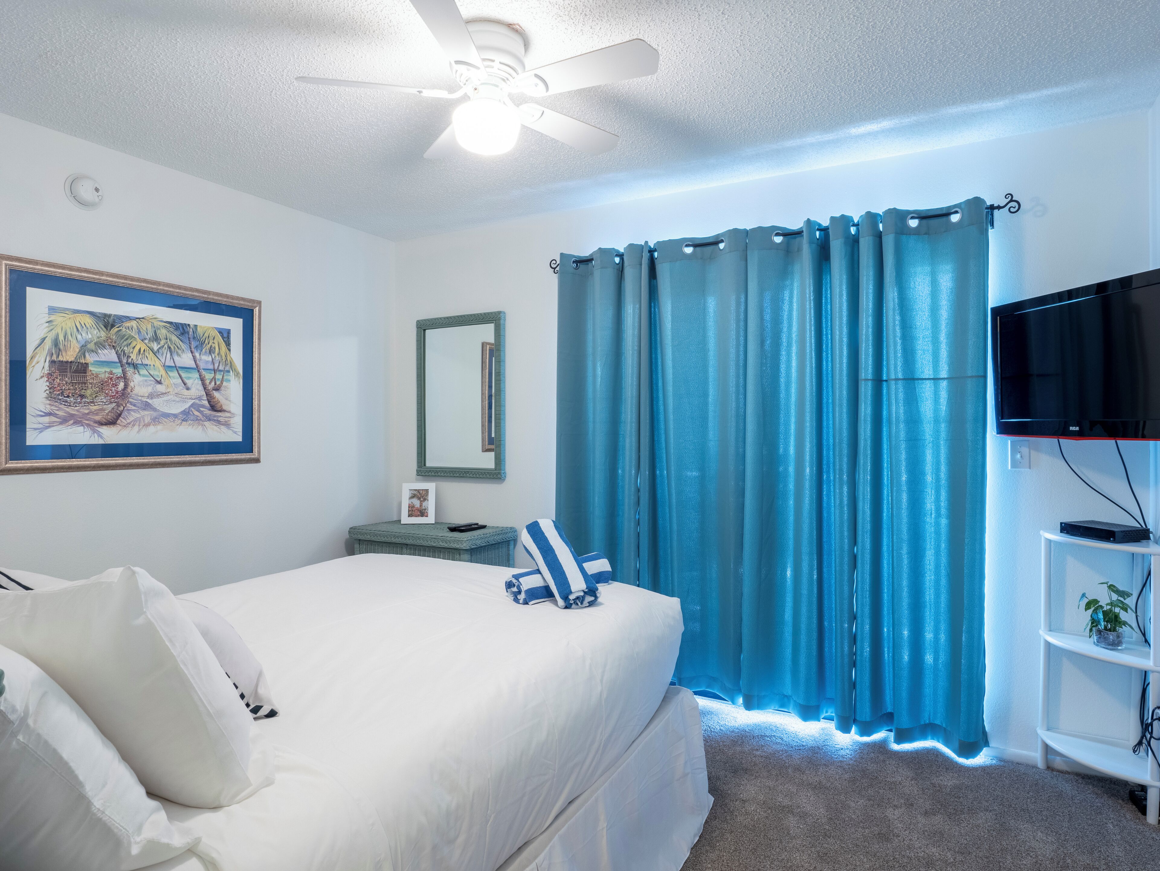 Un dormitorio con cama, ventilador de techo, televisor, espejo y ventana con cortinas azules.