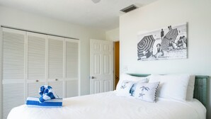 A bedroom with a large bed, white walls, a door with louvered shutters, and a beach-themed artwork on the wall.