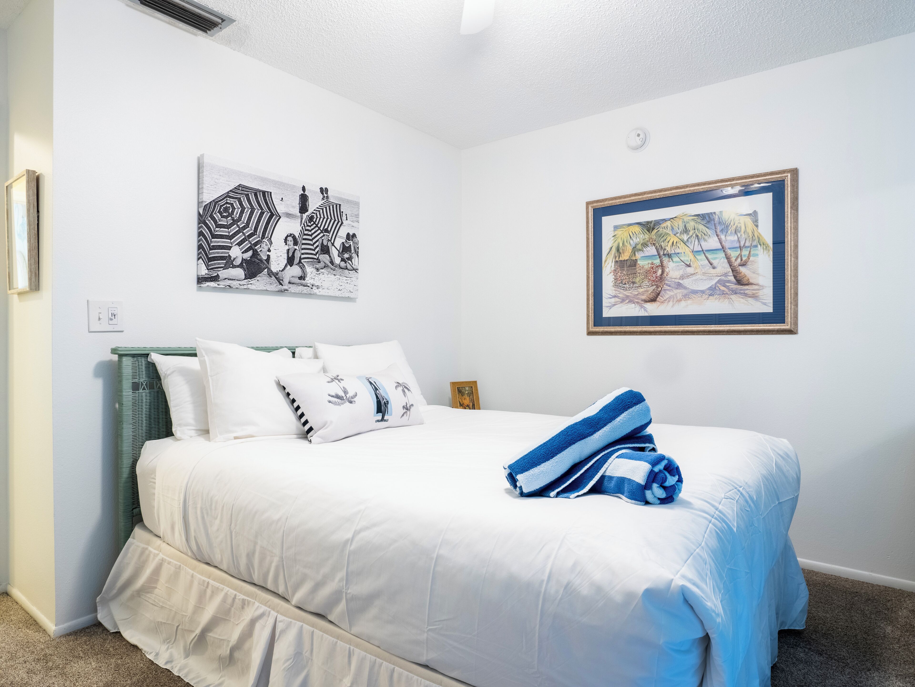 A neatly made bed with white bedding and a blue-striped towel, a green wicker headboard, and two wall-mounted artworks depicting beach scenes.