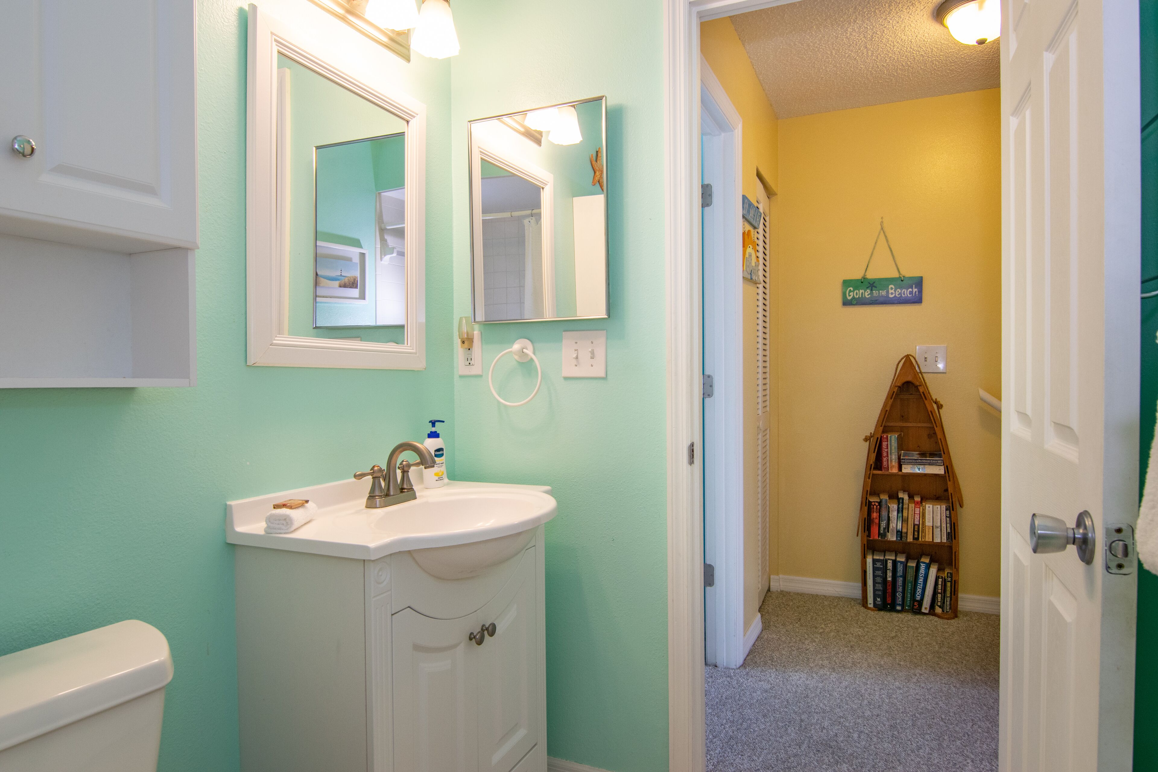 A bathroom with a white sink, two mirrors, and a toilet. There is a bookshelf with books in the background.