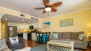 A living room with a sofa, armchair, and coffee table, a kitchen with white cabinets, and a ceiling fan.