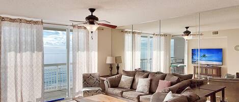 A spacious living room with a large sectional sofa, a coffee table, and a TV. There is a balcony with curtains and a ceiling fan.
