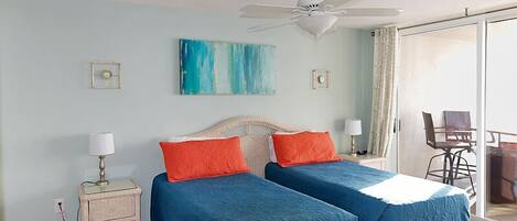 A room with two beds, a ceiling fan, a painting, and a view of the outside.