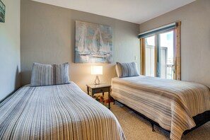 Een hotelkamer met twee bedden, een schilderij van zeilboten, een nachtkastje met een lamp en een raam met uitzicht.