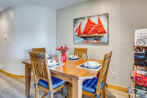 Een eetruimte met een houten tafel gedekt voor vier personen, stoelen met blauwe kussens, een vaas met rode bloemen, en een schilderij van een zeilboot aan de muur.