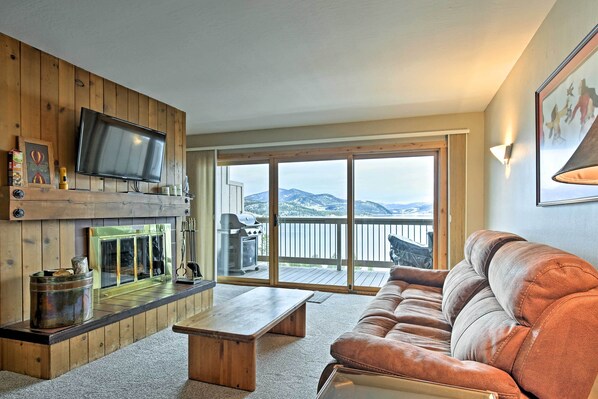 A cozy living room with a wooden wall, a flat-screen TV, a sofa, and a large sliding glass door leading to a balcony with a mountain view. - Mtn-View Condo w/ Hot Tub Access in Dillon! (Dillon)