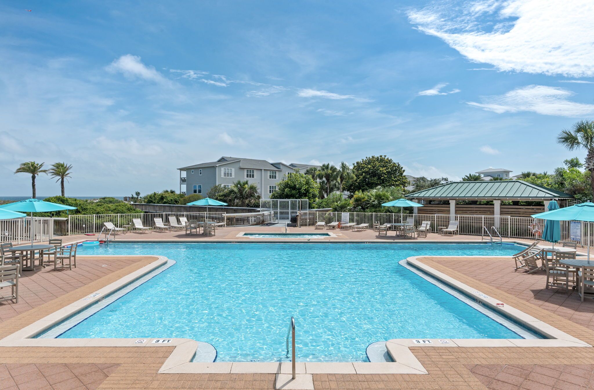 A swimming pool with lounge chairs and umbrellas, surrounded by a fence, with a building in the background.