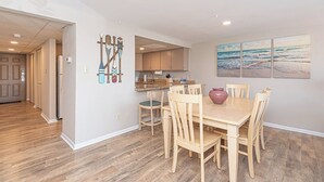 Zona pranzo con tavolo e sedie in legno, cucina sullo sfondo e decorazioni murali a tema spiaggia.