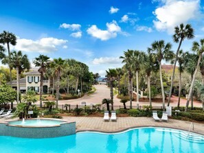 A resort pool area with a clear view of the pool, lounge chairs, palm trees, and a distant building.