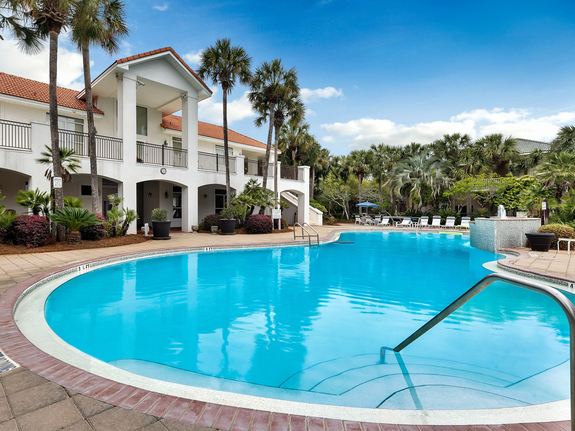 A large, clear swimming pool with a pool ladder, surrounded by palm trees and a multi-story building with balconies.