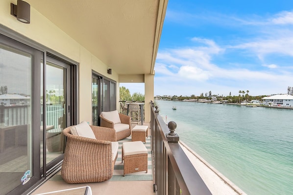 A balcony with wicker furniture, a view of a calm body of water, and a distant shoreline with buildings.
