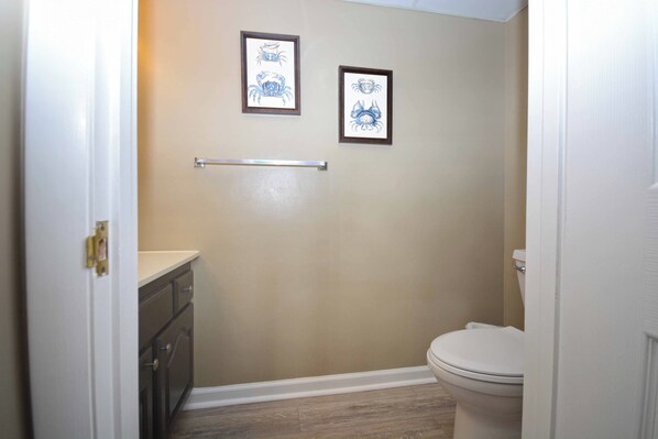 A bathroom with a beige wall, a white toilet, a framed crab artwork, and a metal towel rack. - Spacious, impeccably-styled 1 bedroom oceanfront condo with free WiFi and fabulous view of the ocean just steps to the beach! (Ocean City)