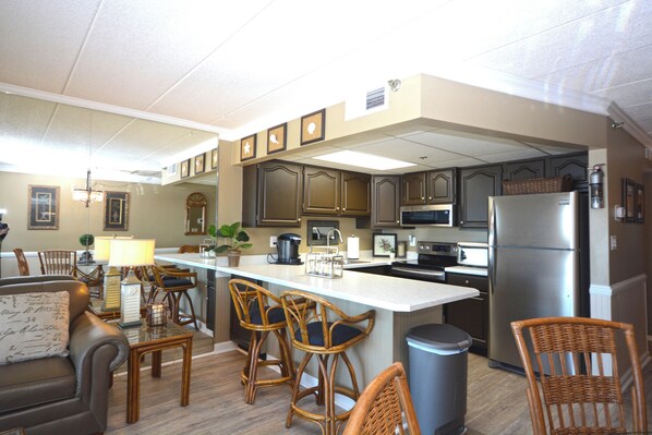A modern kitchen with a central island, bar stools, a refrigerator, and a dining area with a sofa and chairs.