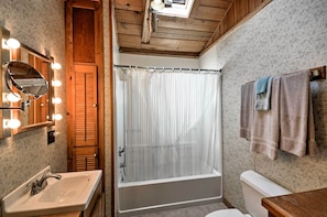 A bathroom with a wooden ceiling, a bathtub, a toilet, a sink, and a mirror with lights.