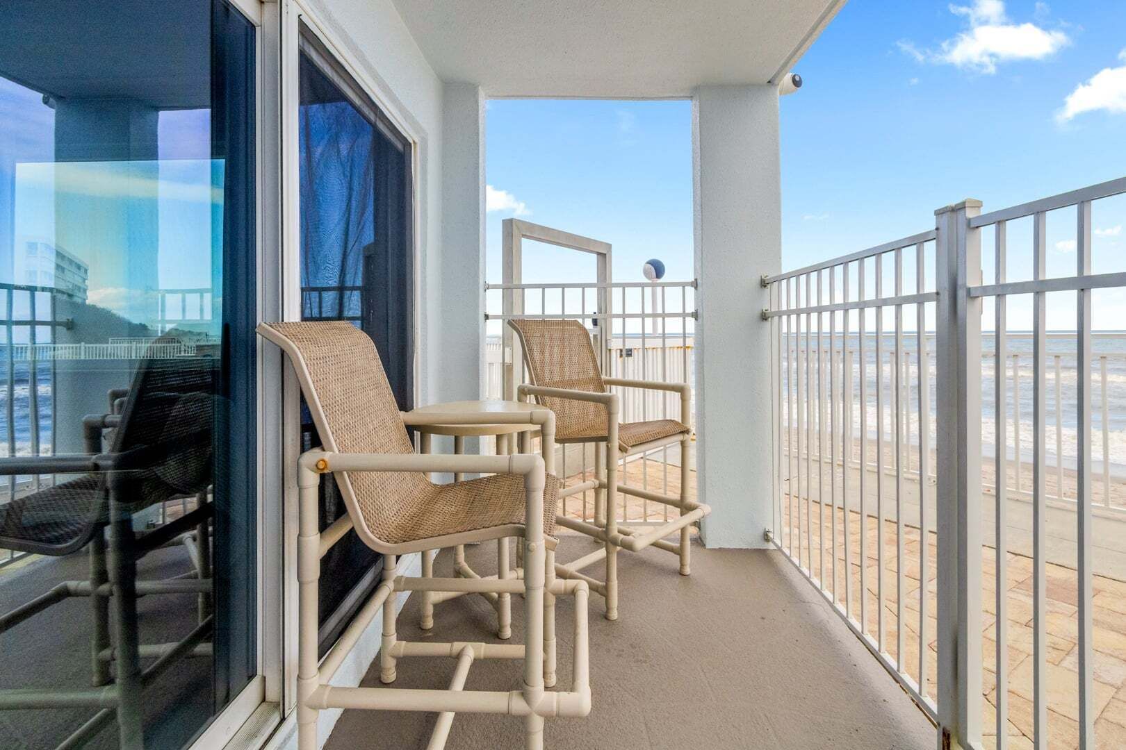 A balcony with two wicker chairs, a glass door, and a white railing overlooking a beach.