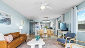 A living room with a brown leather sofa, a white coffee table, and a blue chair. There is a dining area with a white table and chairs, and a kitchen in the background.