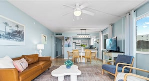 A living room with a brown leather sofa, a white coffee table, and a blue chair. There is a dining area with a white table and chairs, and a kitchen in the background. - Direct Oceanfront Corner Unit with Stunning Views – Just Steps from the Beach! (Satellite Beach)