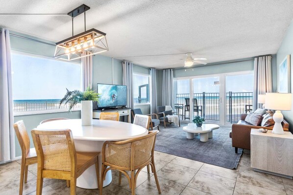 A modern living room with a dining area, a television, and a balcony with a view of the beach.