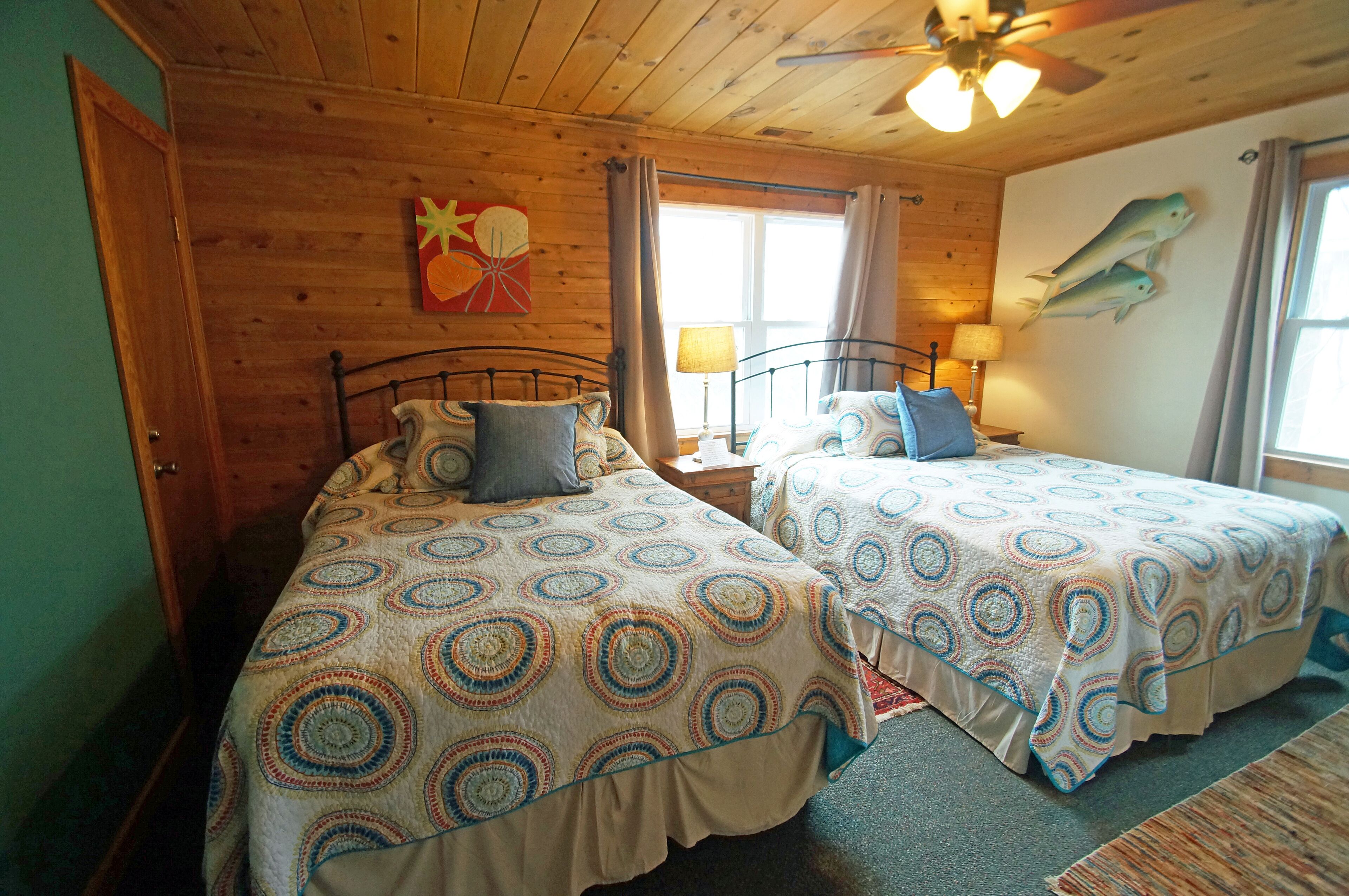 A room with two beds, wooden walls, a ceiling fan, and a window with curtains.