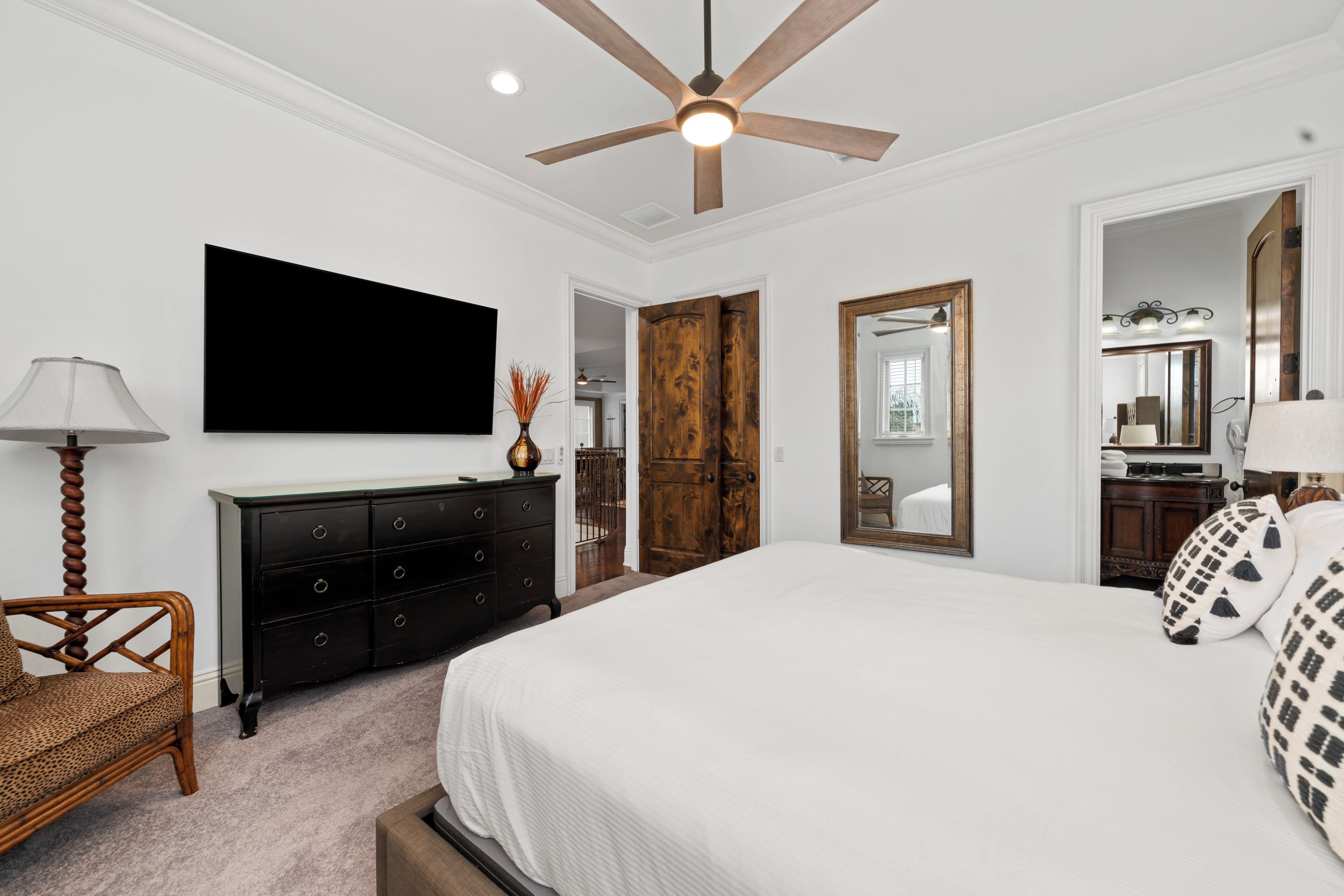 A bedroom with a large bed, a ceiling fan, a TV mounted on the wall, a dresser, and a door leading to another room.