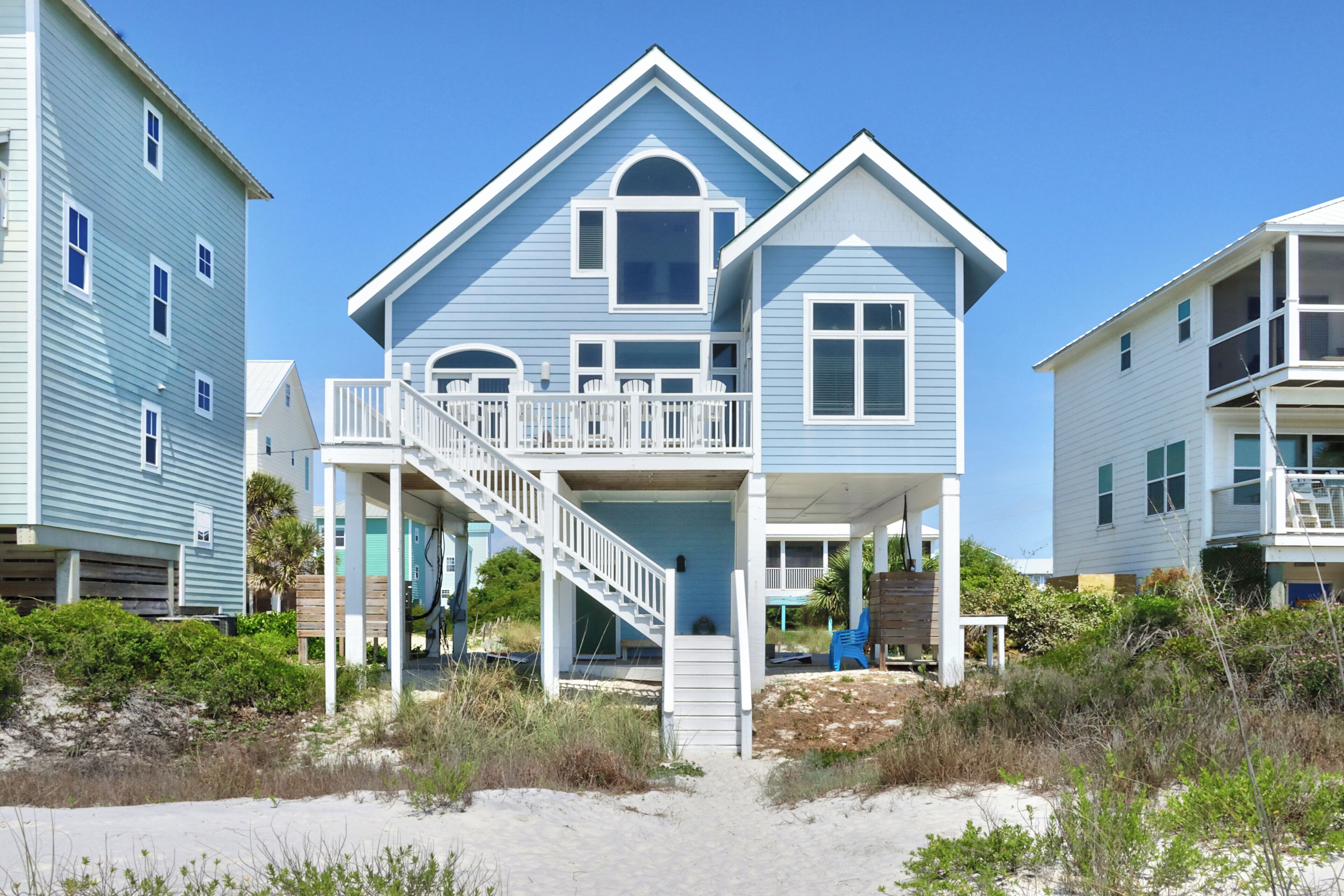 Une maison située en bord de mer, dotée d’un balcon, d’un escalier menant à l’entrée et d’une plage de sable en premier plan.