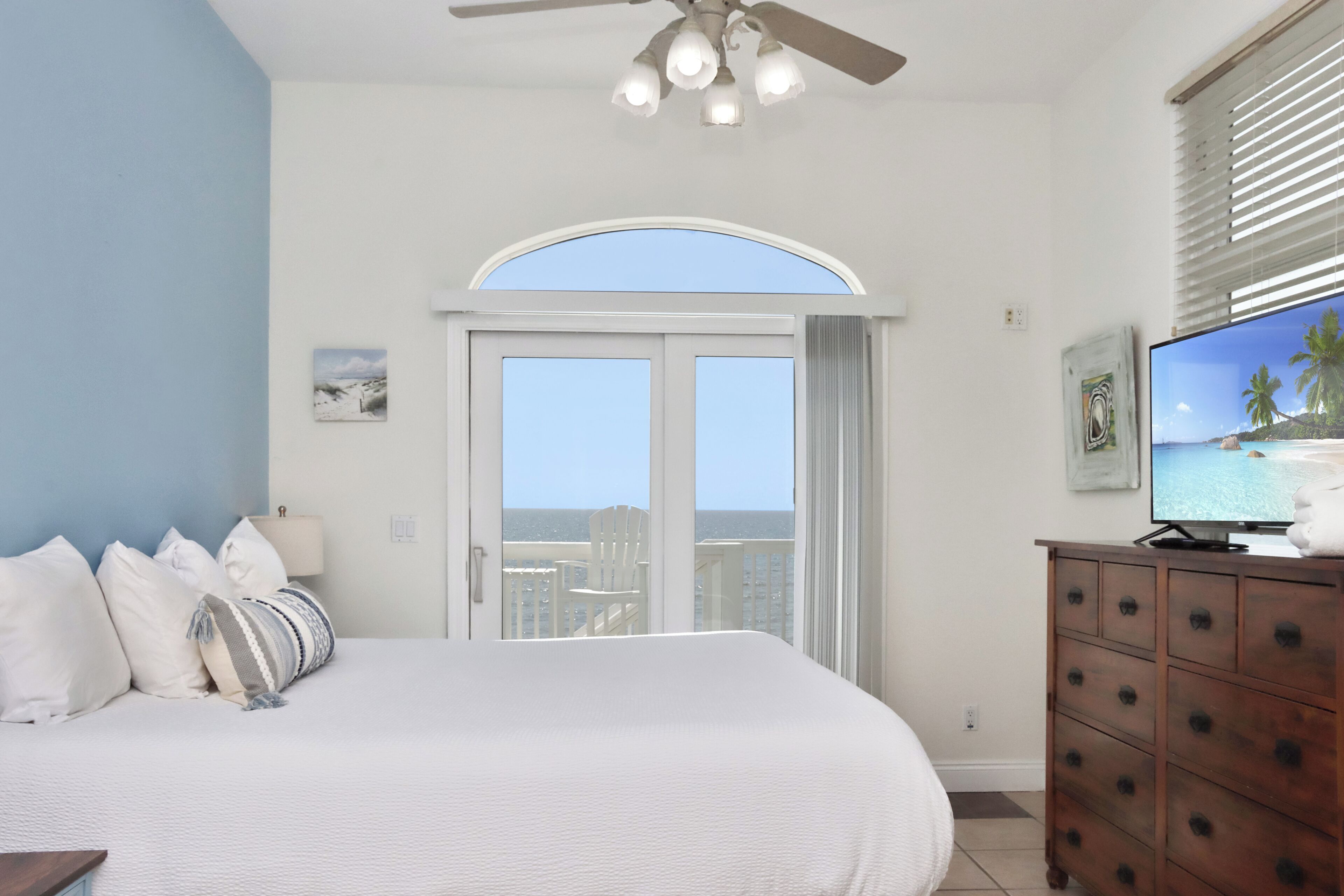 Quarto com cama, cômoda, televisão e vista para o oceano através de uma janela grande.