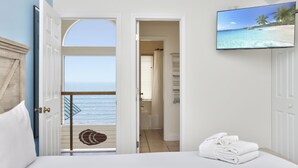 A hotel room with a balcony, a bed with white linens, a television displaying a beach scene, and a view of the ocean. - Sunroom, Quiet Neighborhood, Gulf Front, Private Dune Walkover ~ La Mer (Cape San Blas)