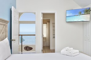 Quarto de hotel com varanda, cama com roupa de cama branca, televisão exibindo uma cena de praia e vista para o mar.