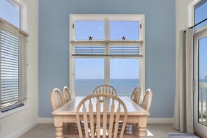 Uma zona de refeições com mesa e cadeiras, vista para o mar através de uma janela e porta para varanda.