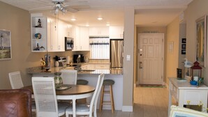 A compact kitchen with white cabinets, a stainless steel refrigerator, and a dining area with white chairs. - Cute 2/2!Direct Oceanfront Complex on the No Drive Beach in NSB! (New Smyrna Beach)