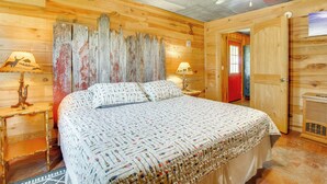 Una habitación tipo cabaña de madera con una cama, dos mesitas de noche con lámparas, un ventilador de techo y una puerta que da a otra habitación.