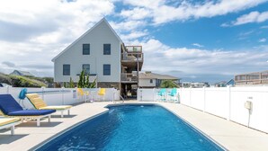 Ein Schwimmbecken mit Liegestühlen, ein mehrstöckiges Gebäude mit Balkon und ein klarer Himmel.
