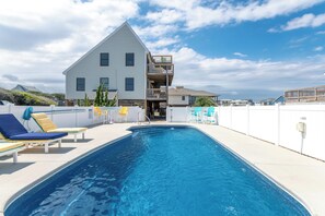 Een zwembad met ligbedden, een gebouw met twee verdiepingen en een balkon, en een heldere lucht.