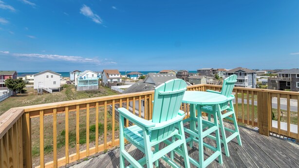 Eine Holzdar Terrasse mit zwei Stühlen und einem Tisch, mit Blick auf einen Strand und eine Wohngegend.