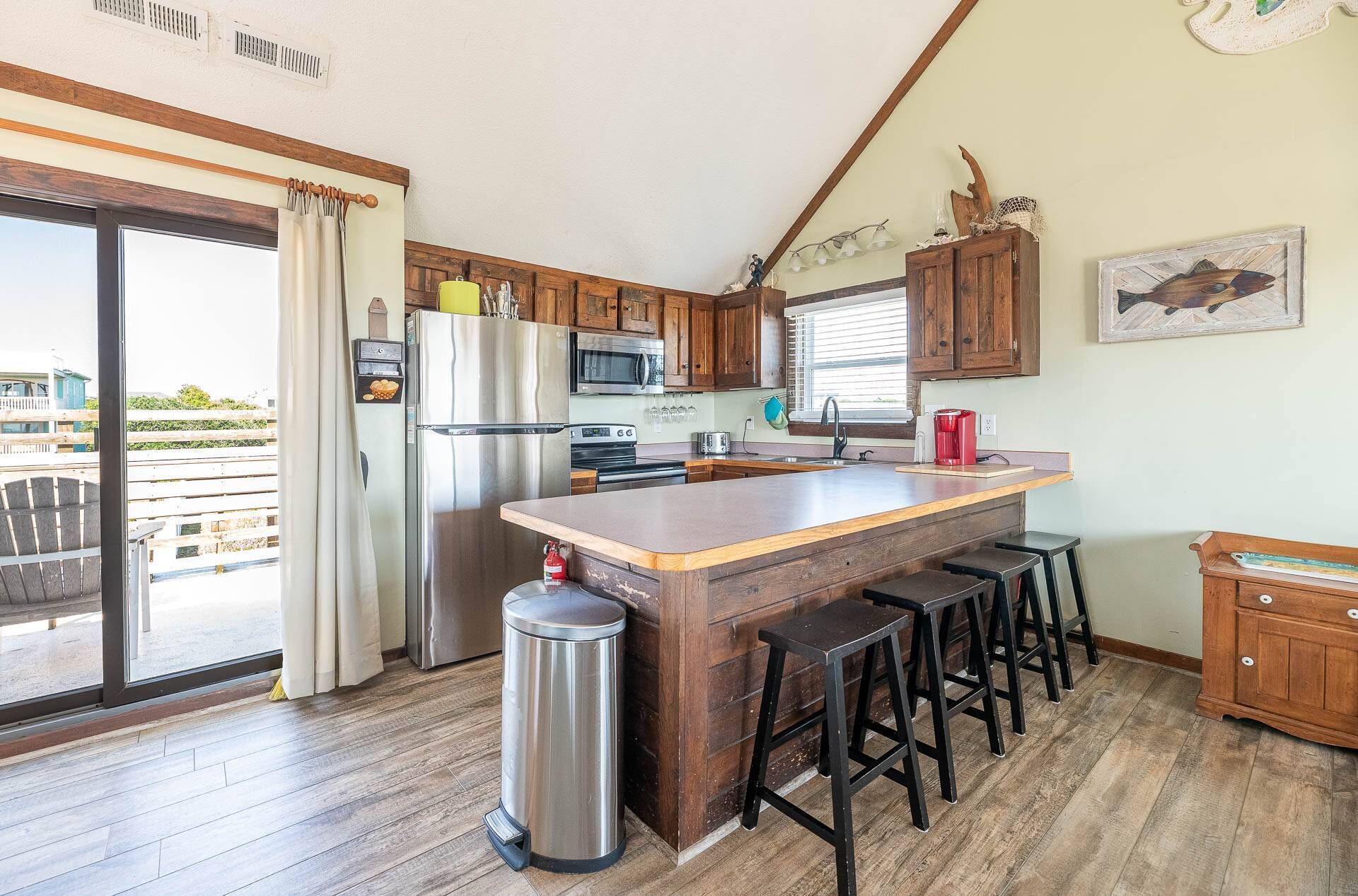 A kitchen with wooden cabinets, a stainless steel refrigerator, a microwave, a sink, and a window with blinds. There are stools and a trash can.