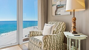 Um quarto com vista para a praia, uma poltrona estampada, uma mesa com abajur e um quadro na parede.