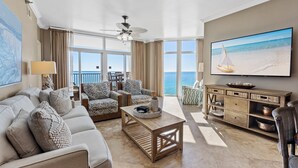 Uma sala de estar ampla, com uma televisão grande, assentos confortáveis, uma mesa de centro e vista para o mar.
